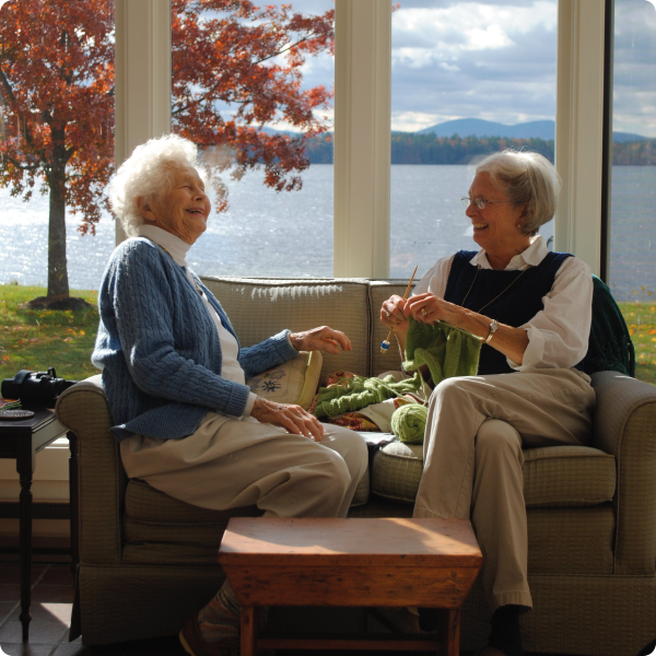 Two elderly women knitting in front of a window laughing